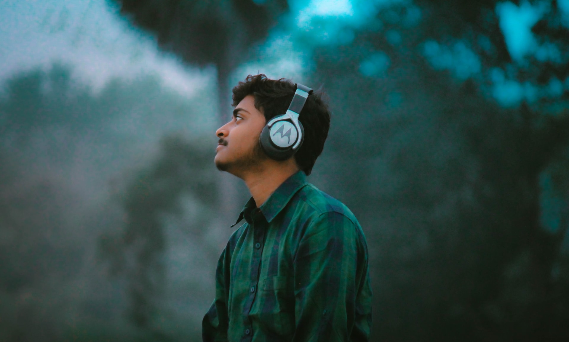 Bild som visar en person i rutig skjorta som bär hörlurar och lyssnar med grönska i bakgrunden.