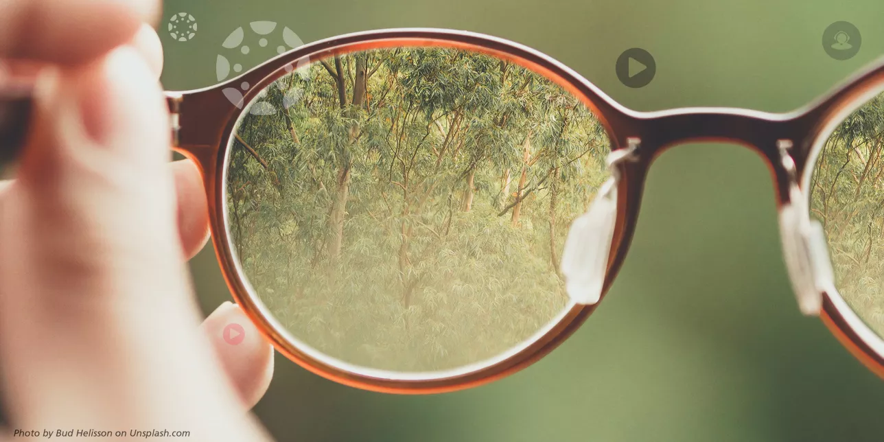Foto av en hand i närbild som håller ett par glasögon med tydligt fokus genom glaset.
