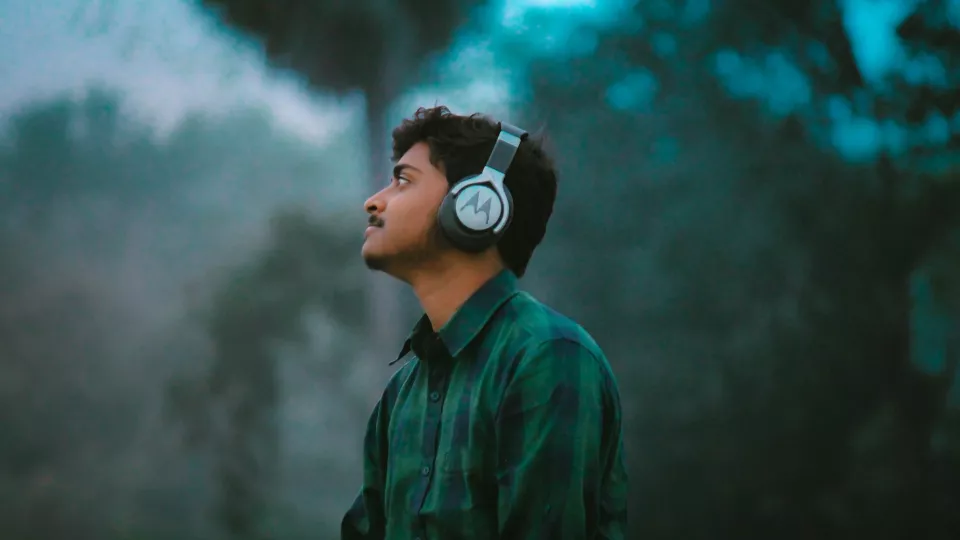 En person i rutig skjorta som bär hörlurar och lyssnar med grönska i bakgrunden.