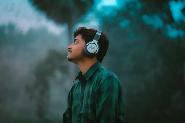 En person i rutig skjorta som bär hörlurar och lyssnar med grönska i bakgrunden.